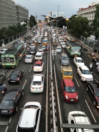 A densely packed urban traffic scene featuring a variety of vehicles, including buses, cars, and motorbikes, moving in both directions on a multi-lane road. Trees and buildings line the sides of the street, with traffic signals and streetlights visible. The atmosphere is busy and congested, typical of metropolitan rush hour.