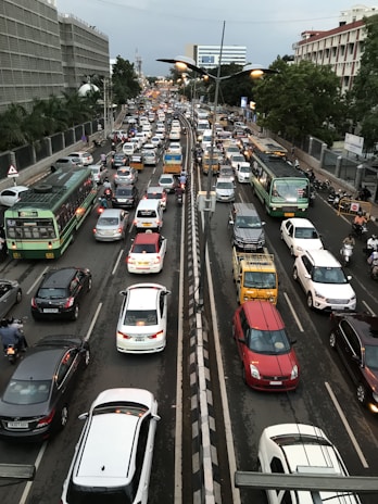 A densely packed urban traffic scene featuring a variety of vehicles, including buses, cars, and motorbikes, moving in both directions on a multi-lane road. Trees and buildings line the sides of the street, with traffic signals and streetlights visible. The atmosphere is busy and congested, typical of metropolitan rush hour.