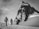 Two climbers are trekking across a snow-covered landscape towards a towering, rugged mountain peak. They are equipped with climbing gear and appear determined as they progress through the icy environment. The scene is monochromatic, giving it a timeless and adventurous feel.
