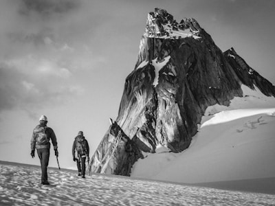 Two climbers are trekking across a snow-covered landscape towards a towering, rugged mountain peak. They are equipped with climbing gear and appear determined as they progress through the icy environment. The scene is monochromatic, giving it a timeless and adventurous feel.