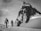 Two climbers are trekking across a snow-covered landscape towards a towering, rugged mountain peak. They are equipped with climbing gear and appear determined as they progress through the icy environment. The scene is monochromatic, giving it a timeless and adventurous feel.