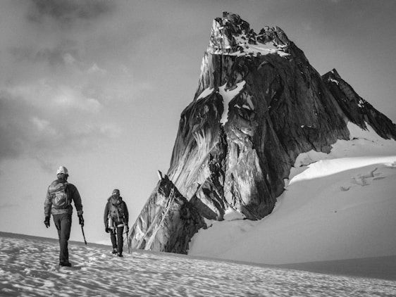 Two climbers are trekking across a snow-covered landscape towards a towering, rugged mountain peak. They are equipped with climbing gear and appear determined as they progress through the icy environment. The scene is monochromatic, giving it a timeless and adventurous feel.