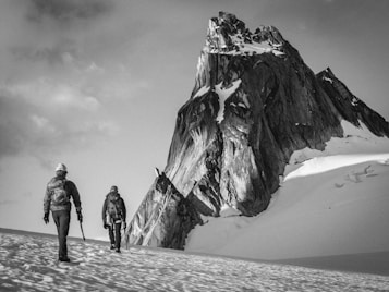 Two climbers are trekking across a snow-covered landscape towards a towering, rugged mountain peak. They are equipped with climbing gear and appear determined as they progress through the icy environment. The scene is monochromatic, giving it a timeless and adventurous feel.