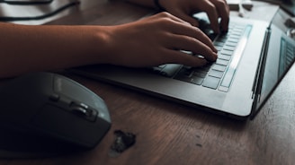 Hands typing on a laptop keyboard, positioned on a wooden desk. A wireless mouse is placed to the left of the laptop.