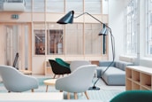 A modern office waiting area features stylish furniture, including contemporary armchairs in shades of teal and gray, arranged around small round tables. A large, curved floor lamp adds a design element, contrasting with the light wood and glass paneling that lines the walls. Natural light streams through large windows, illuminating the space and creating a bright, airy atmosphere.