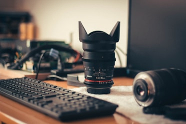 A computer keyboard is placed on a desk alongside a camera lens standing upright, with another lens lying on a tablecloth. In the background, electronic components and a computer monitor are visible.