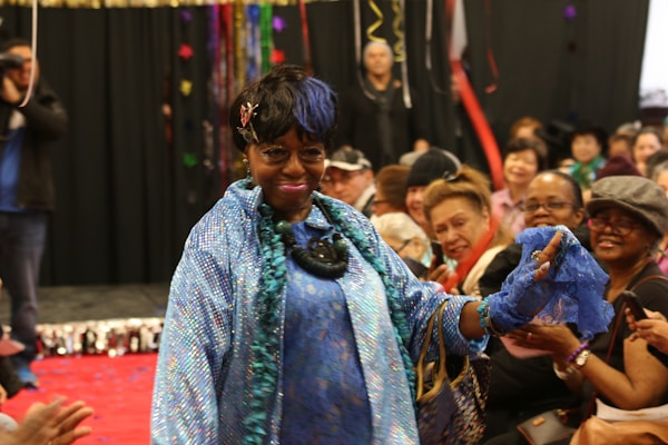 An older woman is confidently walking down a red carpet, adorned in a sparkly blue outfit with matching gloves, surrounded by an audience seated on either side. The audience members appear to be of diverse backgrounds, watching her with smiles and clapping their hands. The setting suggests a festive or celebratory event.