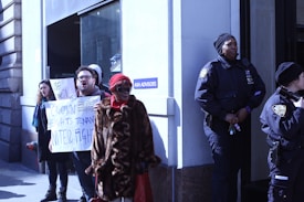 A group of people is standing outside a building, with two police officers observing them. One person is holding a protest sign which reads 'CROWN HEIGHTS TENANT UNITE & FIGHT'. The weather appears cold and some individuals are wearing hats and coats.
