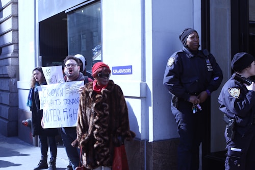 A group of people is standing outside a building, with two police officers observing them. One person is holding a protest sign which reads 'CROWN HEIGHTS TENANT UNITE & FIGHT'. The weather appears cold and some individuals are wearing hats and coats.