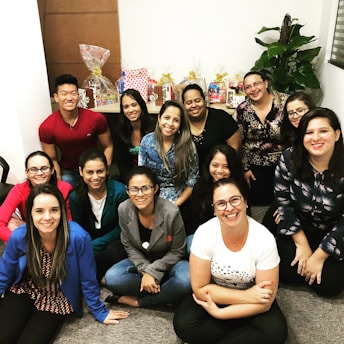 A group of thirteen people, mostly women, are smiling and sitting closely together, suggesting they are part of an informal gathering or celebration. Objects such as gift bags and plants are visible in the background, creating a warm and inviting ambiance.