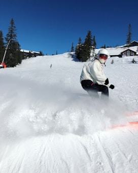 A skier wearing a white jacket, black pants, and a helmet with goggles is seen in mid-action on a snowy slope. Snow is spraying up from the skis, indicating motion. In the background, there are more people skiing and snowboarding downhill, surrounded by snow-covered trees and a few cabin-like buildings under a clear blue sky.
