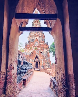 A view through a weathered archway revealing a red-brick temple structure with intricate details. The path leading to the temple is lined with headless Buddha statues on one side. The ancient architectural design and surrounding textures convey a sense of historical and cultural significance.