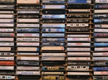 A collection of cassette tapes arranged neatly in stacks. Each tape displays the names of various music albums and artists, featuring diverse genres and time periods. The tapes are weathered and evoke a sense of nostalgia.