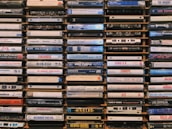 A collection of cassette tapes arranged neatly in stacks. Each tape displays the names of various music albums and artists, featuring diverse genres and time periods. The tapes are weathered and evoke a sense of nostalgia.