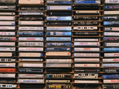 A collection of cassette tapes arranged neatly in stacks. Each tape displays the names of various music albums and artists, featuring diverse genres and time periods. The tapes are weathered and evoke a sense of nostalgia.
