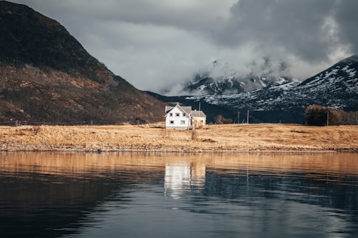 A solitary white house sits on a grassy shoreline, reflected in the calm water of a lake. In the background, mountains are partially covered with snow and shrouded in clouds, creating a dramatic and serene landscape.