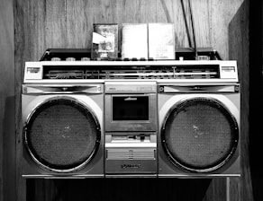 A vintage boombox with two cassette tapes resting on top is set against a wooden background. The boombox features large speakers and a variety of control knobs and tuning indicators, characteristic of retro audio equipment.