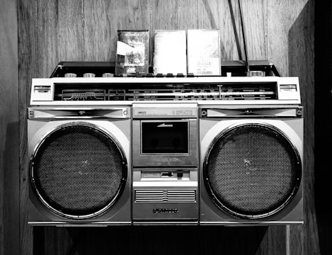 A vintage boombox with two cassette tapes resting on top is set against a wooden background. The boombox features large speakers and a variety of control knobs and tuning indicators, characteristic of retro audio equipment.