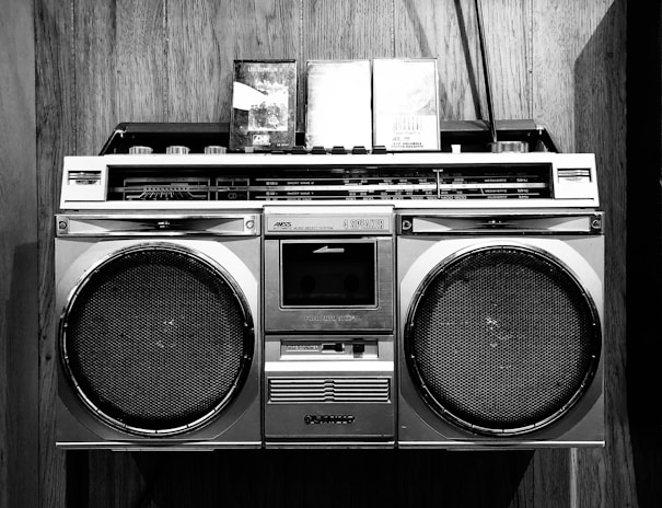 A vintage boombox with two cassette tapes resting on top is set against a wooden background. The boombox features large speakers and a variety of control knobs and tuning indicators, characteristic of retro audio equipment.