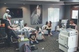 An office environment with multiple people engaged in various tasks. A man wearing a cap is working at a standing desk with a child sitting on his shoulders. Another woman is sitting at a desk using her phone. Two children are on the floor near them, playing or using electronic devices. A woman is seated on a round chair reading to a young girl. A man is working at another desk with a dual-monitor setup near a large printer.