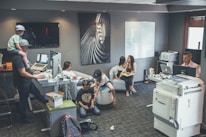 An office environment with multiple people engaged in various tasks. A man wearing a cap is working at a standing desk with a child sitting on his shoulders. Another woman is sitting at a desk using her phone. Two children are on the floor near them, playing or using electronic devices. A woman is seated on a round chair reading to a young girl. A man is working at another desk with a dual-monitor setup near a large printer.