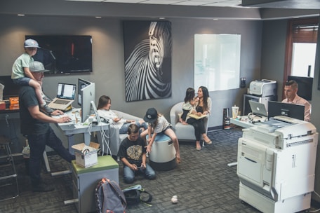 An office environment with multiple people engaged in various tasks. A man wearing a cap is working at a standing desk with a child sitting on his shoulders. Another woman is sitting at a desk using her phone. Two children are on the floor near them, playing or using electronic devices. A woman is seated on a round chair reading to a young girl. A man is working at another desk with a dual-monitor setup near a large printer.