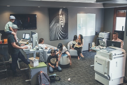 An office environment with multiple people engaged in various tasks. A man wearing a cap is working at a standing desk with a child sitting on his shoulders. Another woman is sitting at a desk using her phone. Two children are on the floor near them, playing or using electronic devices. A woman is seated on a round chair reading to a young girl. A man is working at another desk with a dual-monitor setup near a large printer.