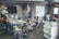 An office environment with multiple people engaged in various tasks. A man wearing a cap is working at a standing desk with a child sitting on his shoulders. Another woman is sitting at a desk using her phone. Two children are on the floor near them, playing or using electronic devices. A woman is seated on a round chair reading to a young girl. A man is working at another desk with a dual-monitor setup near a large printer.