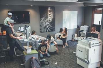 An office environment with multiple people engaged in various tasks. A man wearing a cap is working at a standing desk with a child sitting on his shoulders. Another woman is sitting at a desk using her phone. Two children are on the floor near them, playing or using electronic devices. A woman is seated on a round chair reading to a young girl. A man is working at another desk with a dual-monitor setup near a large printer.