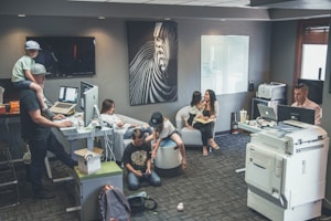 An office environment with multiple people engaged in various tasks. A man wearing a cap is working at a standing desk with a child sitting on his shoulders. Another woman is sitting at a desk using her phone. Two children are on the floor near them, playing or using electronic devices. A woman is seated on a round chair reading to a young girl. A man is working at another desk with a dual-monitor setup near a large printer.
