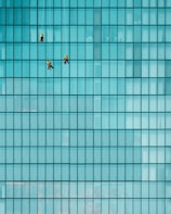 Three workers are suspended on ropes cleaning the windows of a large glass building, creating a pattern with the grid of the building's panes.
