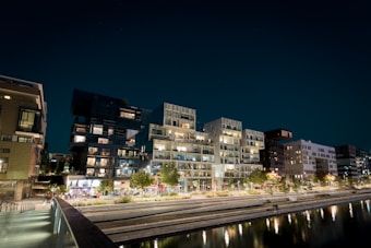 A modern urban residential area at night, featuring a row of stylish, illuminated apartment buildings. The structures have multiple balconies with lights on, highlighting their architectural details. In front of the buildings, there are well-maintained green spaces with trees and a walkway beside a canal that reflects the lights from the buildings.