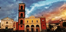 A colorful, historic town square featuring three distinct buildings, each with unique architectural styles. The central building has a yellow facade with arched windows and a clock tower on the left. The sky is dramatic with deep blue and orange hues, adding a vibrant atmosphere.