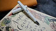 A white fountain pen rests diagonally on a book titled 'Angela Carter's Book of Fairy Tales.' The book features whimsical illustrations of mythical creatures and intricate designs. The background includes a subtle texture with a leather-like object in brown color.