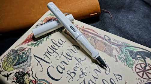 A white fountain pen rests diagonally on a book titled 'Angela Carter's Book of Fairy Tales.' The book features whimsical illustrations of mythical creatures and intricate designs. The background includes a subtle texture with a leather-like object in brown color.