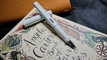 A white fountain pen rests diagonally on a book titled 'Angela Carter's Book of Fairy Tales.' The book features whimsical illustrations of mythical creatures and intricate designs. The background includes a subtle texture with a leather-like object in brown color.