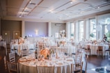 A beautifully decorated banquet hall features round tables adorned with floral centerpieces and elegant table settings, set against a backdrop of soft lighting and large windows. The room has a spacious, airy feel, enhanced by high ceilings and ample natural light coming in through the windows.