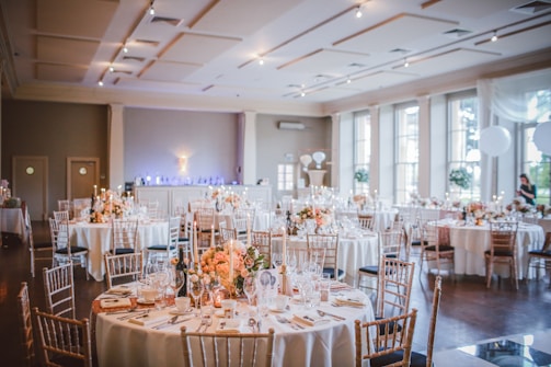 A beautifully decorated banquet hall features round tables adorned with floral centerpieces and elegant table settings, set against a backdrop of soft lighting and large windows. The room has a spacious, airy feel, enhanced by high ceilings and ample natural light coming in through the windows.