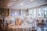 A beautifully decorated banquet hall features round tables adorned with floral centerpieces and elegant table settings, set against a backdrop of soft lighting and large windows. The room has a spacious, airy feel, enhanced by high ceilings and ample natural light coming in through the windows.
