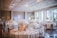 A beautifully decorated banquet hall features round tables adorned with floral centerpieces and elegant table settings, set against a backdrop of soft lighting and large windows. The room has a spacious, airy feel, enhanced by high ceilings and ample natural light coming in through the windows.