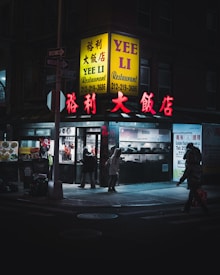 A dimly lit street corner features a Chinese restaurant with bright yellow and red signage that reads 'YEE LI Restaurant'. The windows display hanging roasted meats and inside, figures are seen. People in winter clothing with hoods are passing by on the sidewalk, adding a sense of early evening or night.