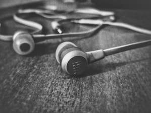 A pair of earphones lies on a textured wooden surface. The earphones are shown in close-up, with one earpiece prominently in focus while the wires and the second earpiece appear blurred in the background, creating a sense of depth.