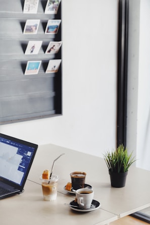 A clean tabletop with a laptop displaying a screen, two glasses of coffee, a small dessert, a plastic cup with a beverage, and a small potted plant. On the wall, there is a display of hanging Polaroid-style photos.