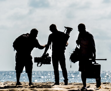 silhouette of men holding camera standing on sand near body of water during daytime