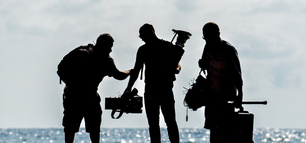 silhouette of men holding camera standing on sand near body of water during daytime