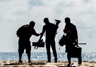 silhouette of men holding camera standing on sand near body of water during daytime