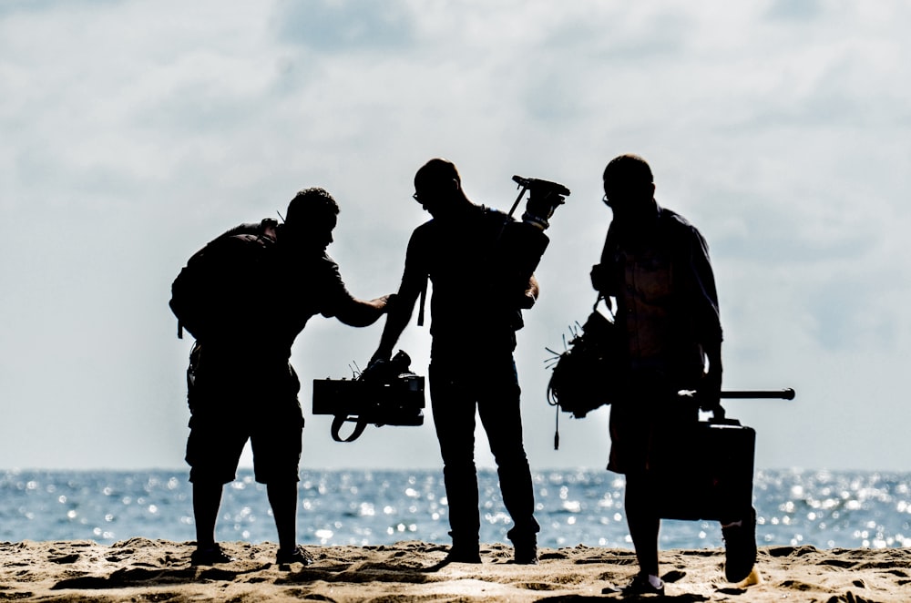 silhouette of men holding camera standing on sand near body of water during daytime