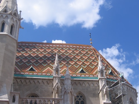 The image features a segment of a building's roof with intricate, colorful tile patterns and ornate stone spires. The roof tiles are arranged in geometric designs, including diamonds and chevrons, with vibrant colors like orange, green, and yellow. Several spires with detailed carvings extend upward along the edges of the roof. A section of scaffolding is visible on the right side, suggesting maintenance or construction work. A clear blue sky with a few scattered clouds forms the backdrop.