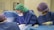 Several medical professionals are engaged in a surgical procedure. They are wearing surgical gowns, masks, caps, and gloves for hygiene and safety. The environment appears to be a sterile operating room with medical equipment around them.