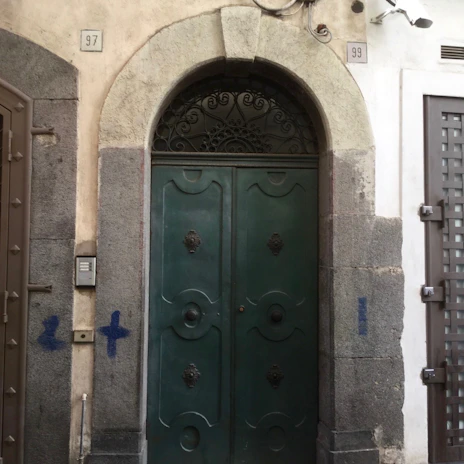 A large, ornate green door set within an archway with intricate metalwork above. The building facade features textured stonework and what appears to be a security camera above. The doors are framed by weathered stone blocks, and there are small blue markings on the stone.
